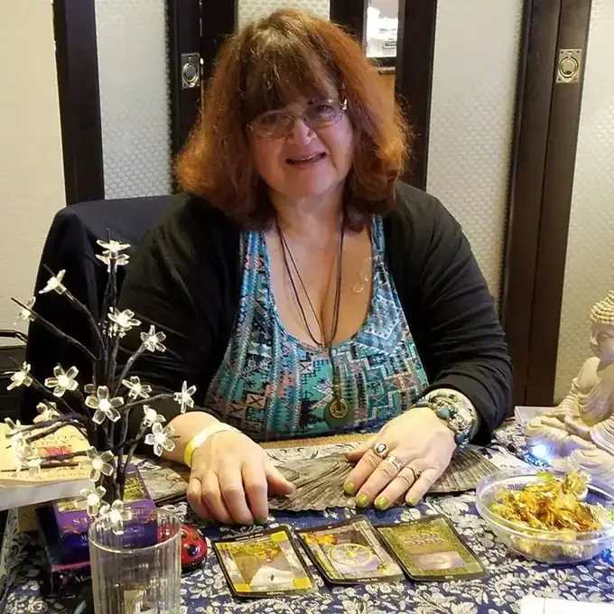 Rev. Berta Kay smiling at her tarot reading table with cards and a lit flower lamp